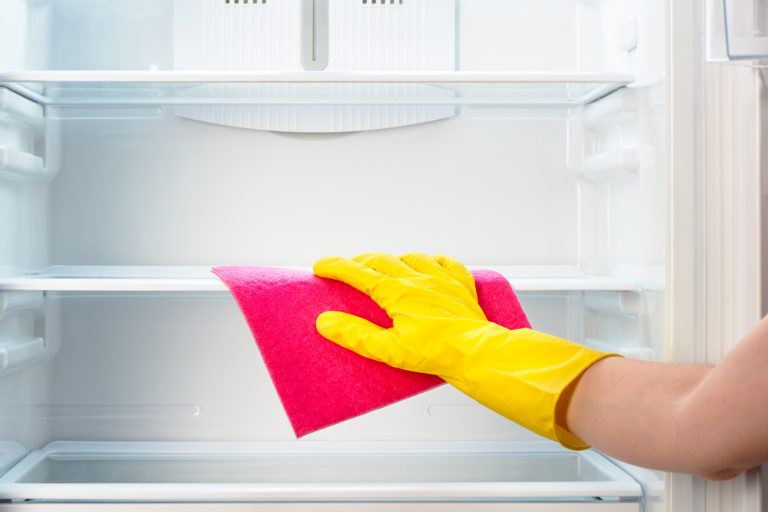 A person wiping down the inside of an empty refrigerator with a pink cloth.