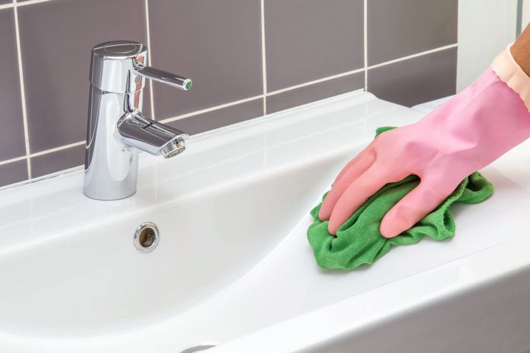 A gloved hand wiping down a bathroom sink with a green microfiber cloth.