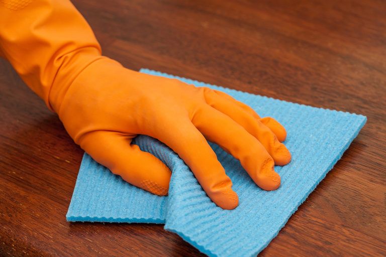 A gloved hand cleans a wooden piece of furniture with a blue rag.