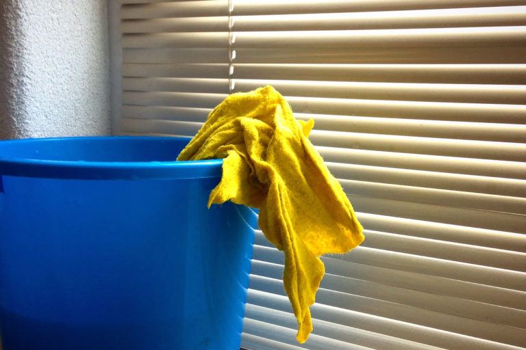 A blue bucket with a yellow rag sticking out sitting in front of closed blinds.