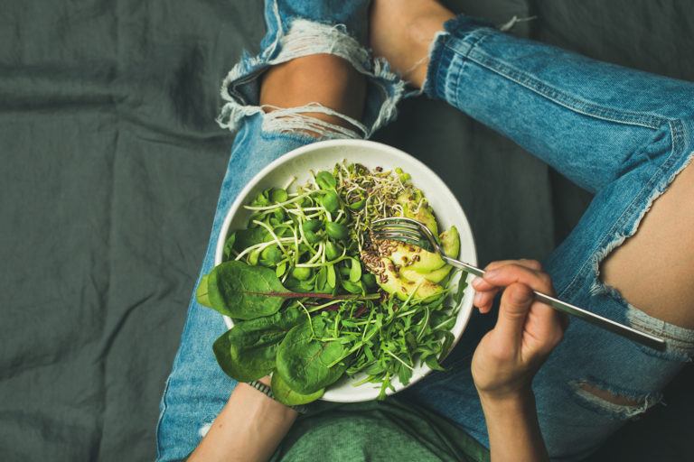 A person in jeans eating a large bowl of greens and avocado.