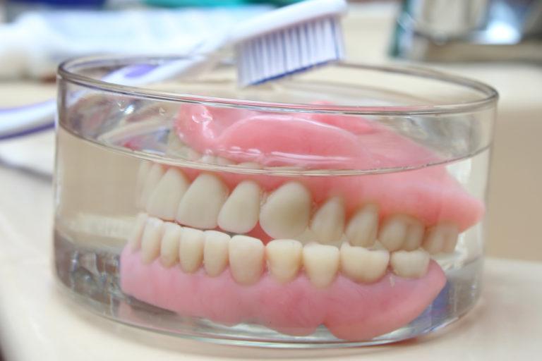 Dentures sitting in a glass jar with a toothbrush laying on the side of the jar.